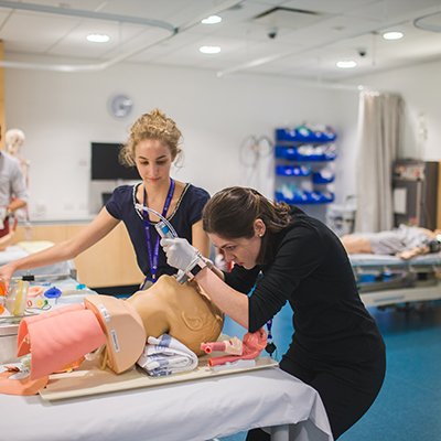 Med students practicing intubating with a dummy on a stretcher. She is bent over the head, while another woman watches